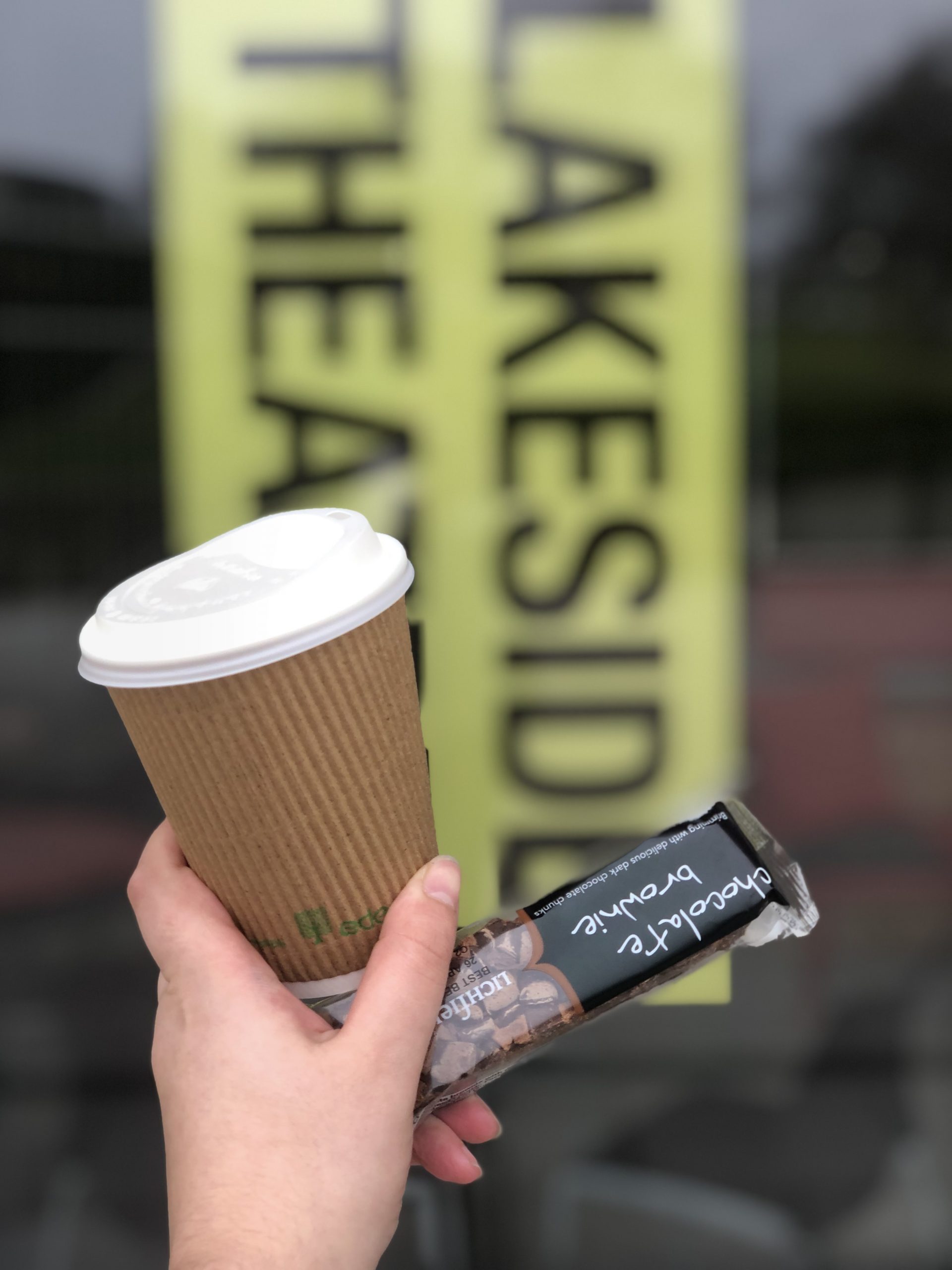 A hand holding a coffee cup and a brownie outside the Lakeside Theatre cafe.