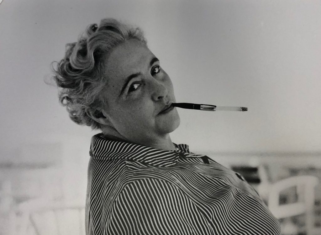 A black and white photo of Margery Allingham looking absolutely fabulous with her coiffed hair and peering over her shoulder while balancing a cigarette holder with lit cigarette between two fingers,