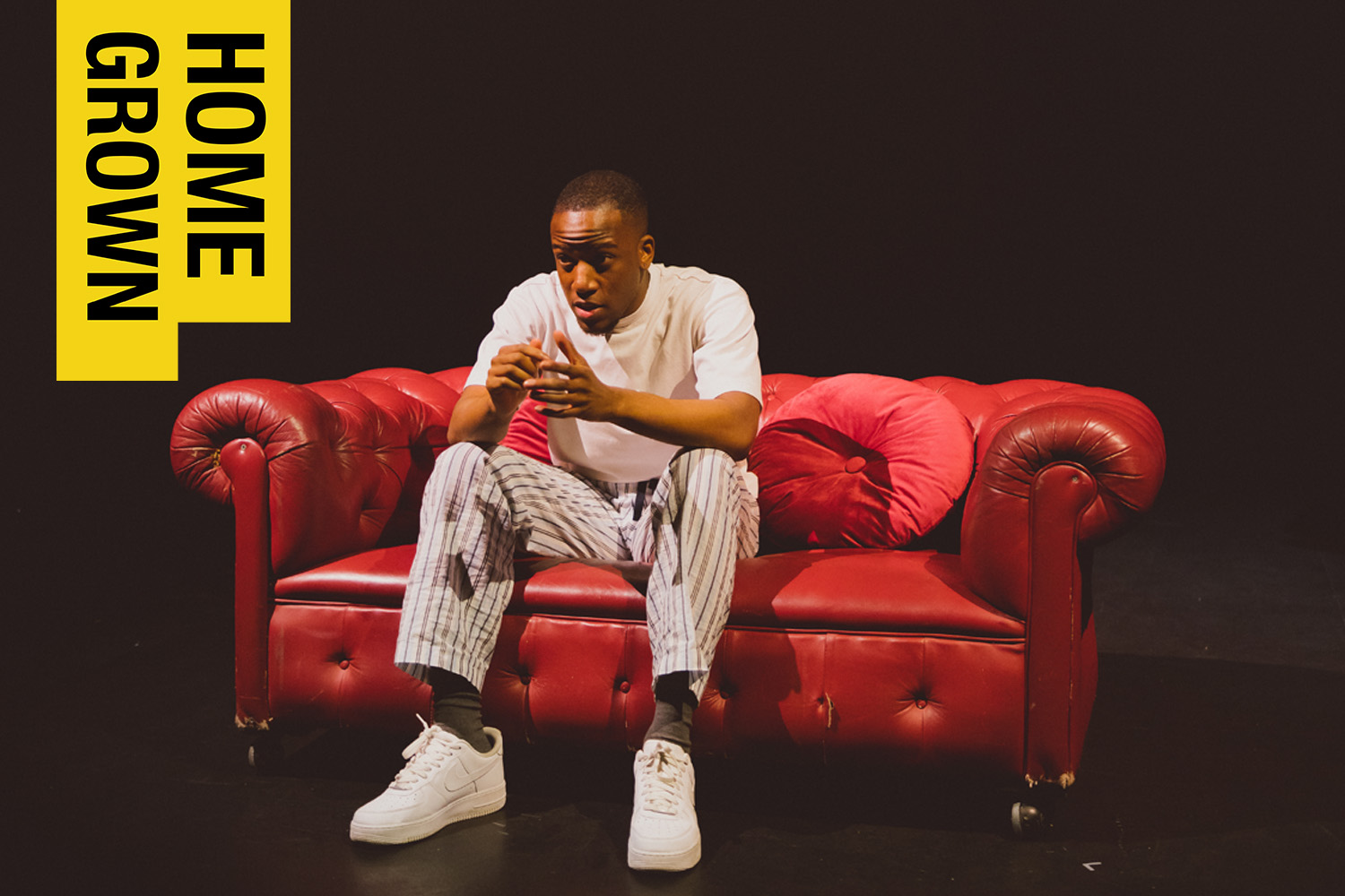 Lakeside Theatre Homegown. A young black man in a light coloured tee-shirt and trousers sits on a red leather couch on a dark stage.