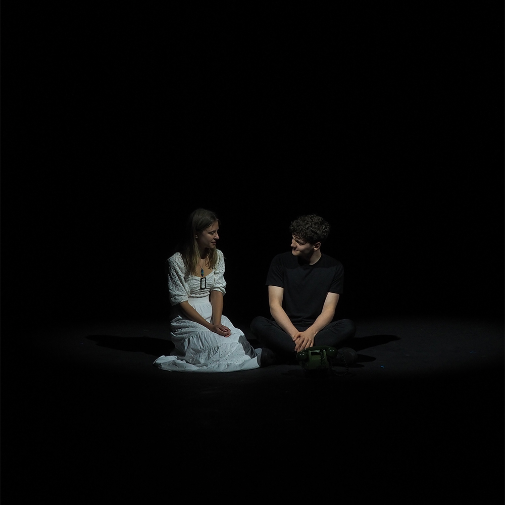 A young woman dressed in white and a young man dressed in black, sit on an empty dark stage with a spotlight slightly illuminating them.