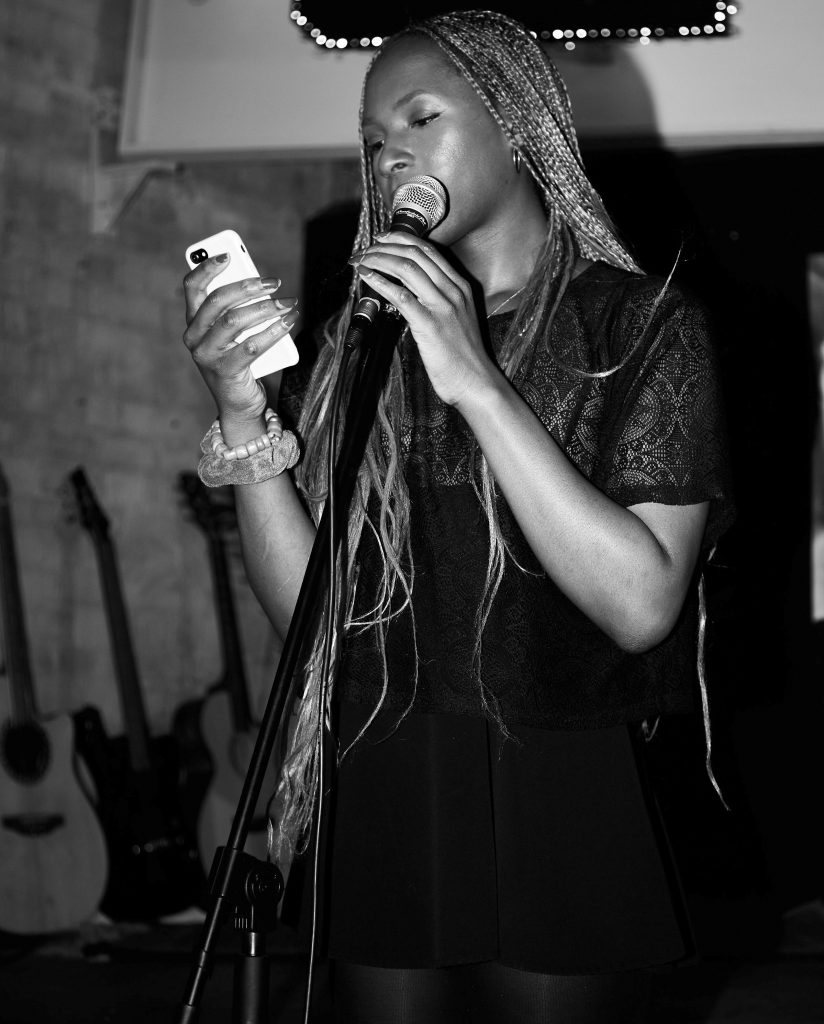 A black and white photo of Chloe Filani, a black, queer poet, artist, performer, and writer. She stands on a stage with a microphone reading from a smartphone screen with her long braids flowing over her shoulder.