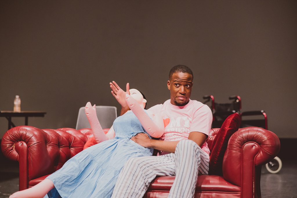 A young black ma sits on a red sofa with a sex doll dressed in a blue dress.
