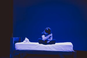 a performance from Launch Festival 2023 where a young woman is sat cross legged on a bed on a dimly lit in blue stage