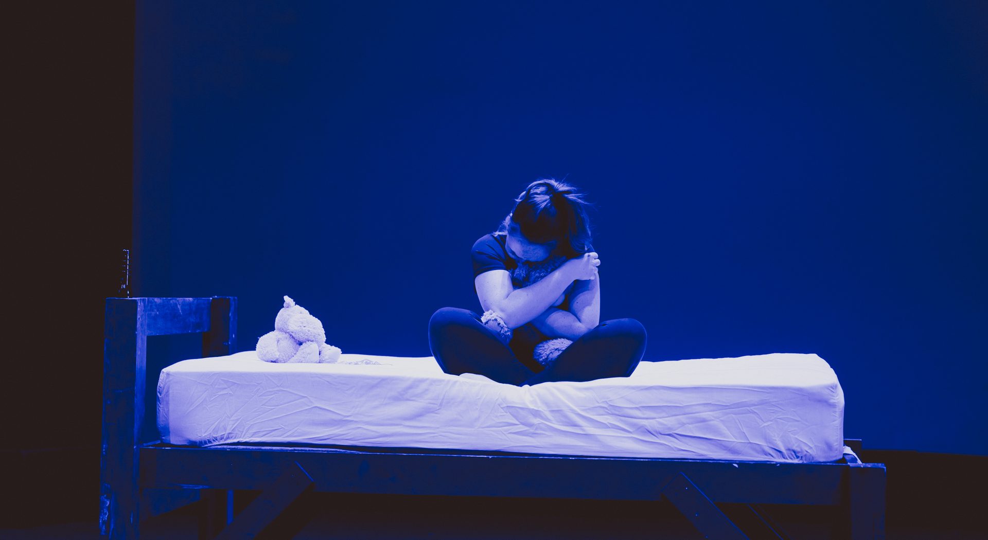 a performance from Launch Festival 2023 where a young woman is sat cross legged on a bed on a dimly lit in blue stage