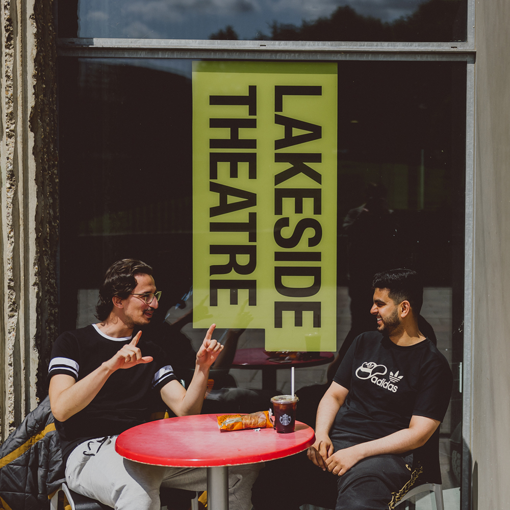 Two young men sit at an outside table ourside the Lakeside Theatre with the LAkeside Theatre branding on the window behind them