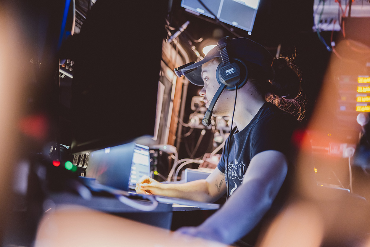 A member of staff from the Lakeside Theatre technical team, working in the tech booth backstage. They're wearing a headset and looking at various controls.