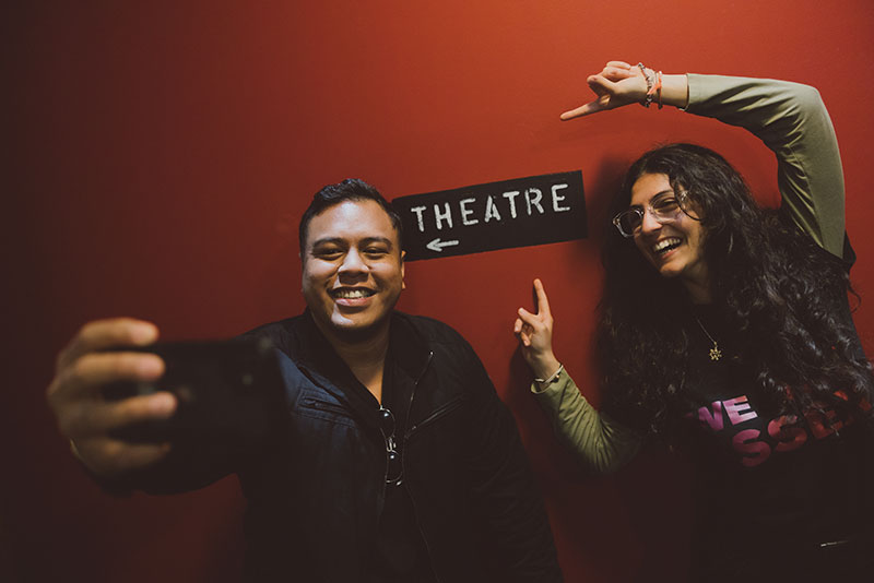 Two smiling students taking a selfie and pointing to the nearby 'theatre' sign in the entrance to the Lakeside Theatre.