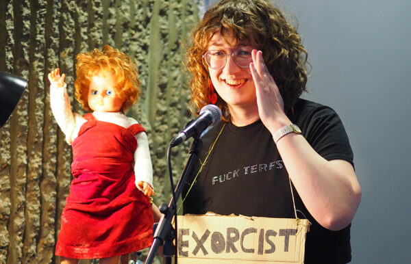 Alice K Stephens performing into a microphone (and holding up a doll) at the Lakeside Theatre Café.