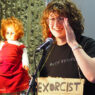 Alice K Stephens performing into a microphone (and holding up a doll) at the Lakeside Theatre Café.